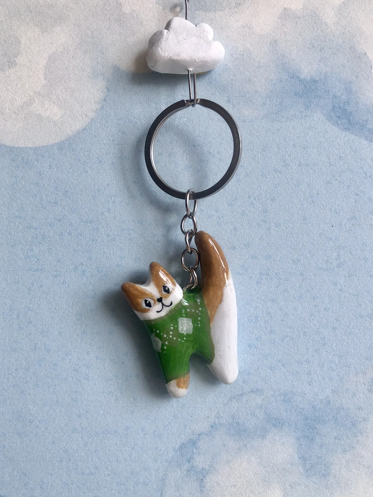 Llavero de gatito naranjo con sweater verde, colgando frente a un cielo con nubes