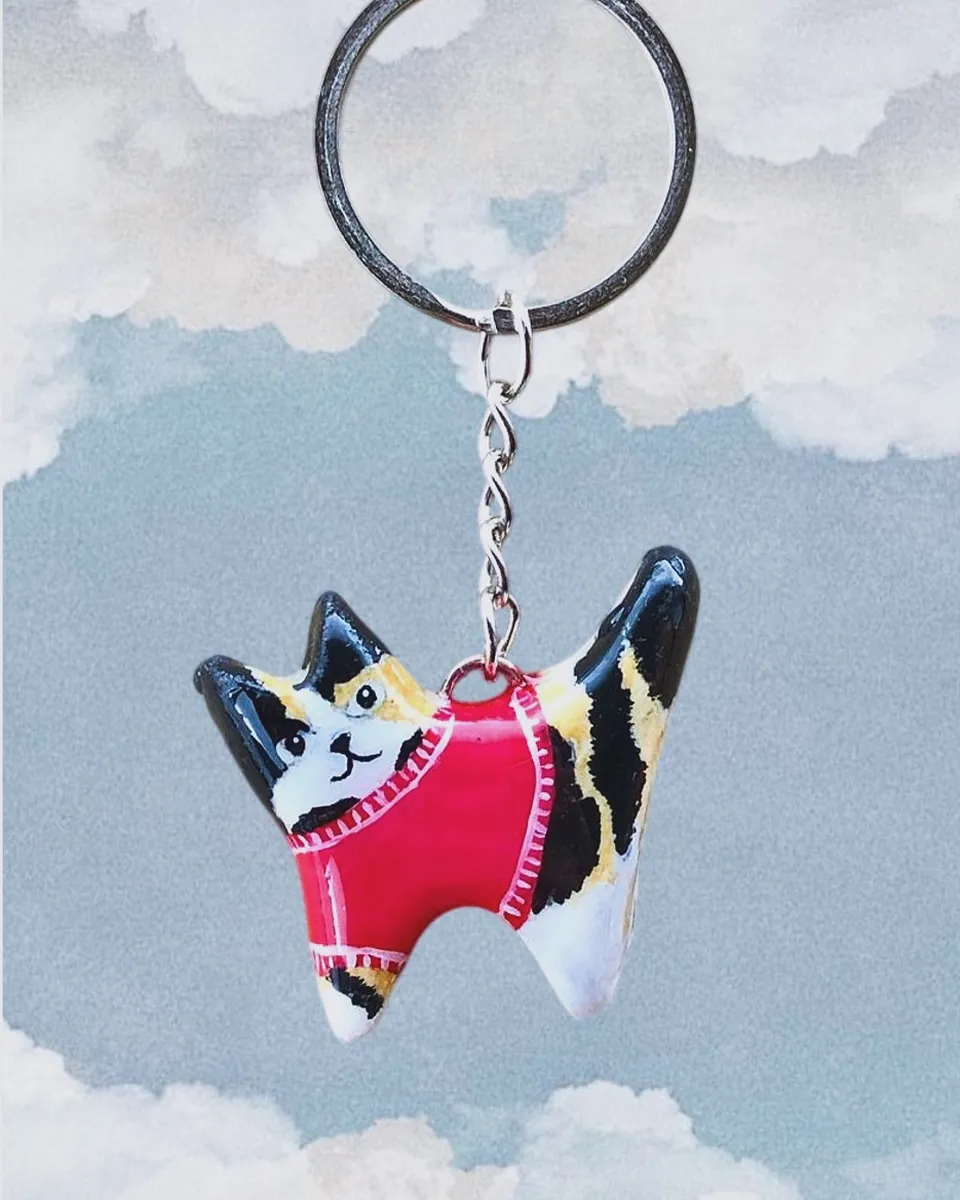Llavero de gatito calico con sweater fucsia frente a un cielo con nubes