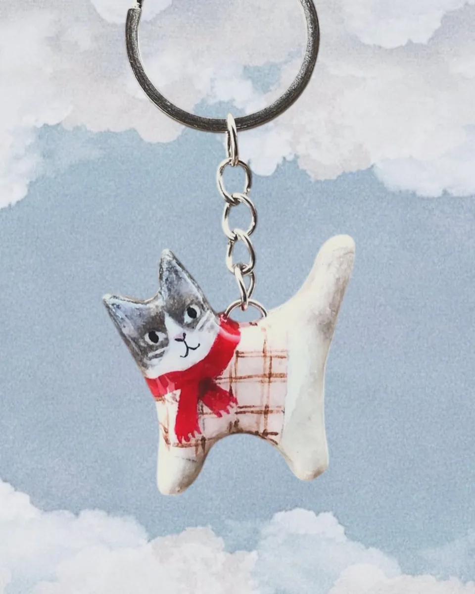 Llavero de gatito gris y blanco con bufanda roja frente a un cielo con nubes