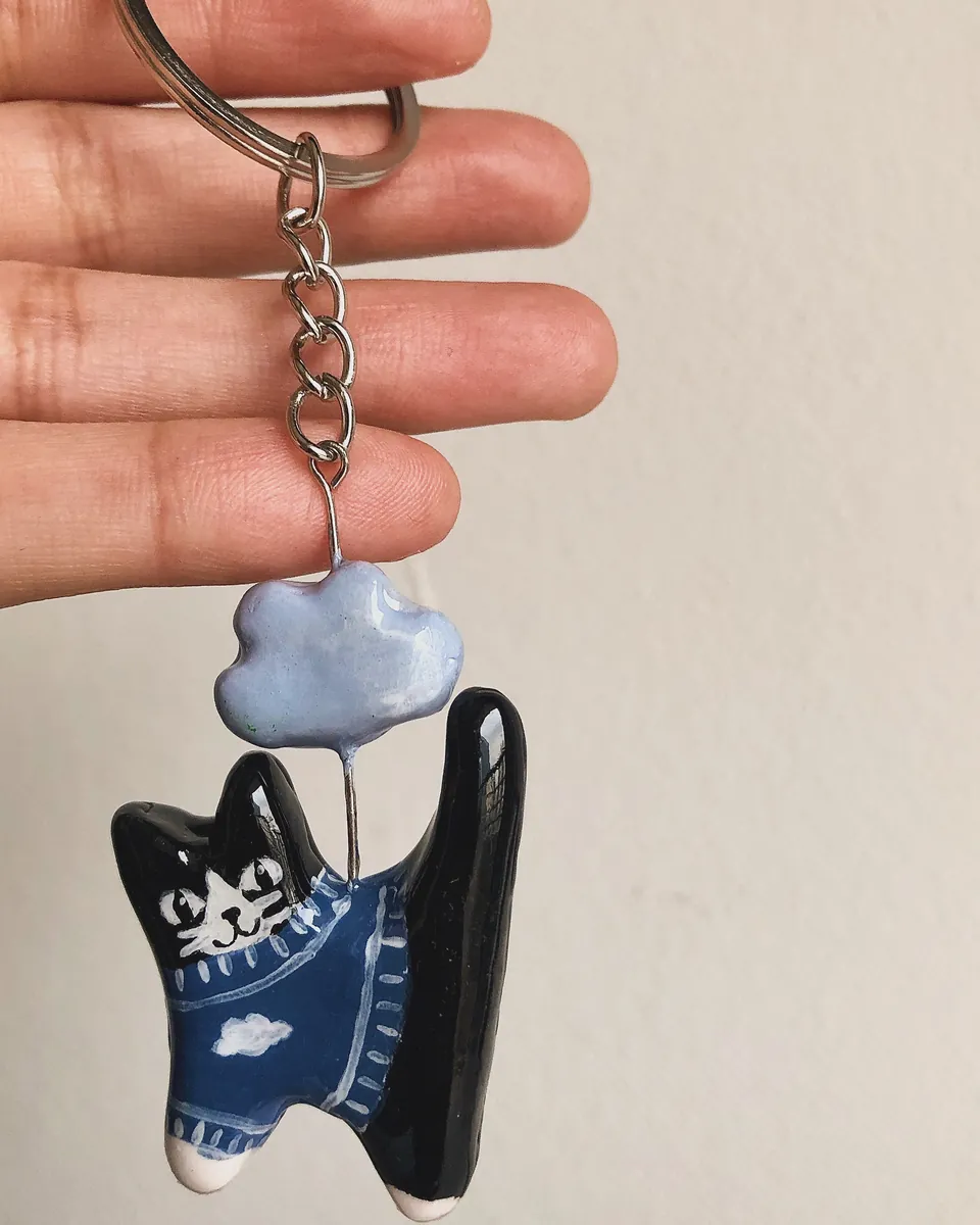 Llavero de gatito blanco y negro con sweater azul colgando de una nube celeste