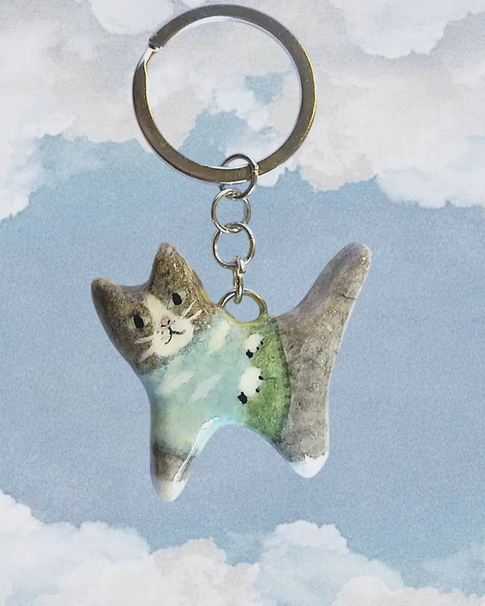 Llavero de gatito gris con sweater verde agua de ovejitas frente a un cielo con nubes