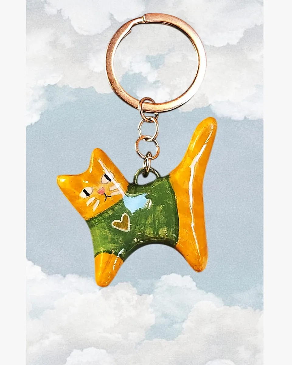 Llavero de gatito naranjo con sweater verde, colgando frente a un cielo con nubes