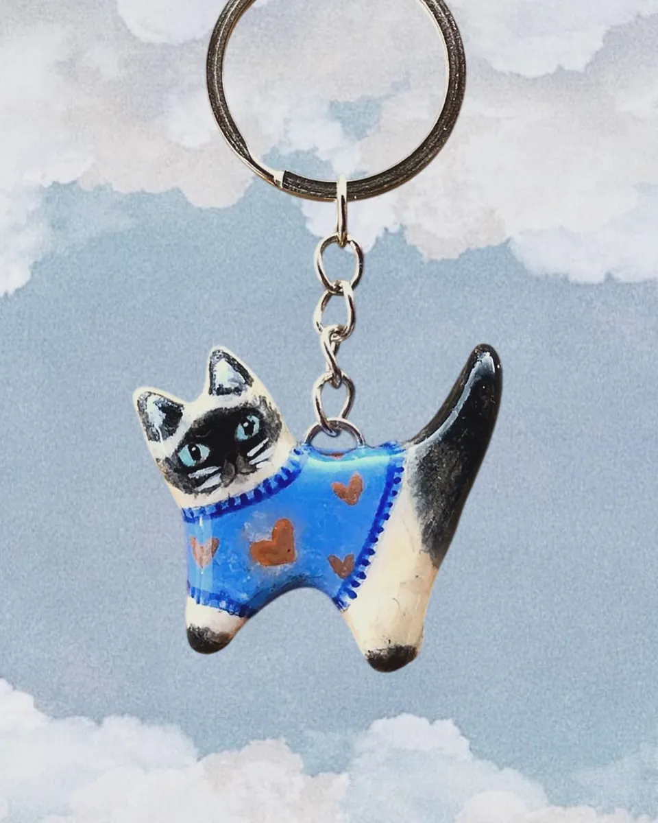 Llavero de gatito siamés con sweater azul de corazones frente a un cielo con nubes